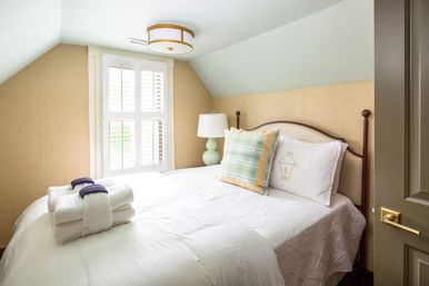 Cozy sunlit attic bedroom with sloped ceiling, white quilted bed, plaid accent pillow, mint bedside lamp, stacked towels and plantation-shutter window.