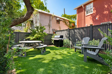 Sunny fenced urban backyard with artificial turf, round picnic table, two gray Adirondack chairs, a gas grill, and shade from a large tree.