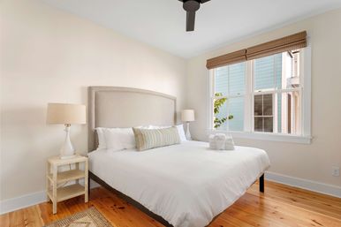 Sunny minimalist bedroom with white bedding, beige upholstered headboard, striped lumbar pillow, wooden nightstand and lamp, hardwood floors, large window with woven shades and ceiling fan.