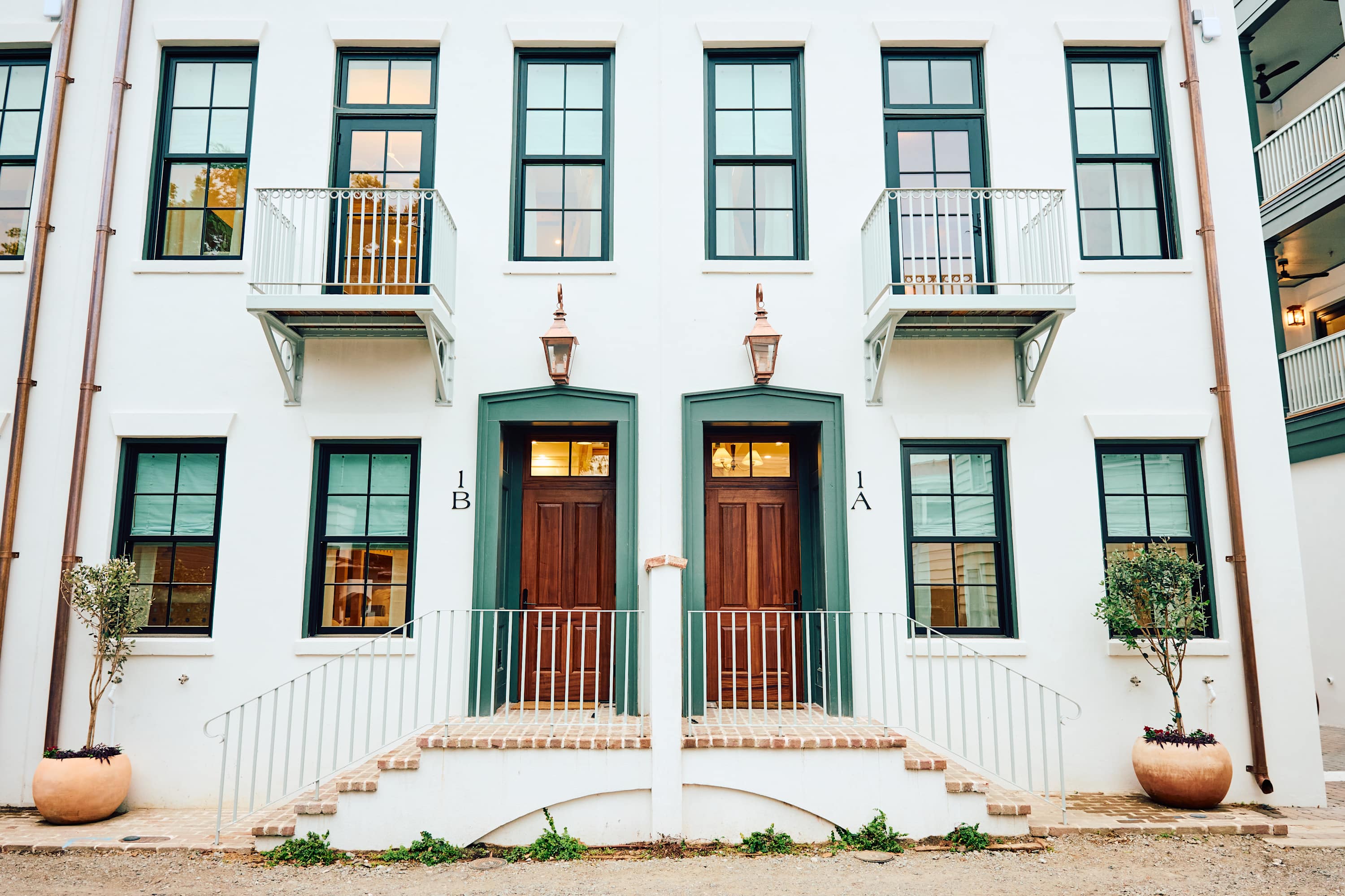 Charming white stucco duplex façade with green-trim windows, twin wooden doors labeled 1A and 1B, small iron balconies, copper lanterns, and potted trees.