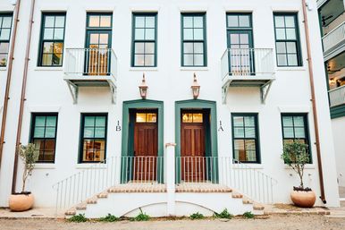 Charming white stucco duplex façade with green-trim windows, twin wooden doors labeled 1A and 1B, small iron balconies, copper lanterns, and potted trees.