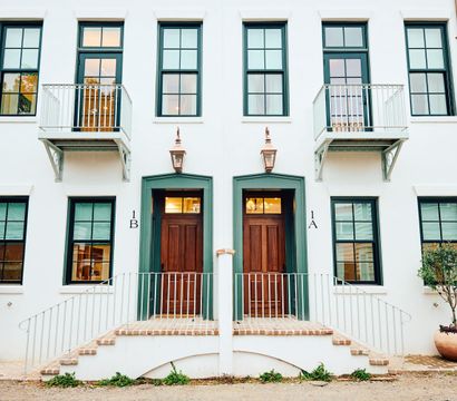 Charming white stucco duplex façade with green-trim windows, twin wooden doors labeled 1A and 1B, small iron balconies, copper lanterns, and potted trees.