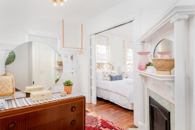 Sunlit boho bedroom interior with mid-century wooden dresser and large round mirror, white fireplace mantel, woven baskets and plants, patterned rug, hardwood floors, and bed with rattan headboard.