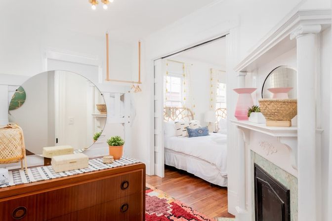 Sunlit boho bedroom interior with mid-century wooden dresser and large round mirror, white fireplace mantel, woven baskets and plants, patterned rug, hardwood floors, and bed with rattan headboard.
