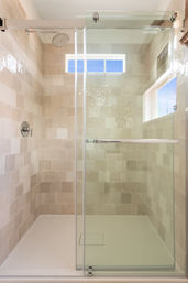 Sunlit modern walk-in shower with beige square tiles, frameless sliding glass door and chrome rain showerhead in a home bathroom alcove.