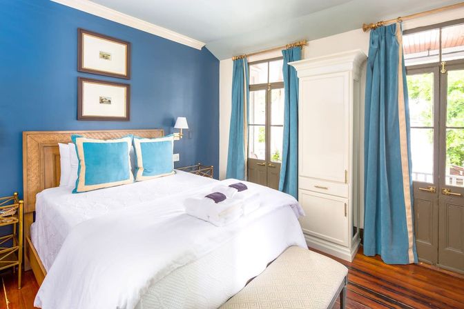 Sunlit cozy guest bedroom with blue accent wall, white bedding, turquoise pillows and curtains, hardwood floors and French doors.