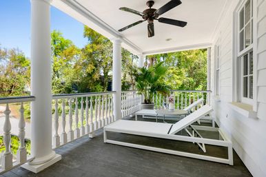 Sunlit white front porch with decorative balustrade and ceiling fan, two white chaise lounges and small table with flowers beside a large potted palm, overlooking a leafy green yard.