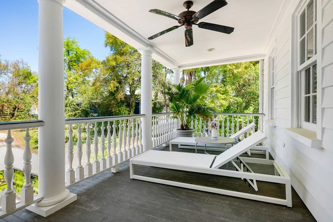 Sunlit white front porch with decorative balustrade and ceiling fan, two white chaise lounges and small table with flowers beside a large potted palm, overlooking a leafy green yard.