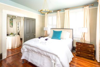 Sunlit bedroom interior with plush white bedding and teal accent pillow, warm hardwood floors, wooden nightstands, cream curtains, gray door and brass chandelier.