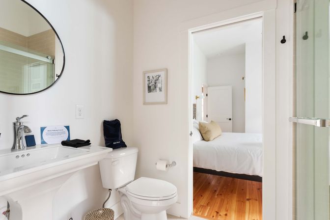 Bright minimalist ensuite bathroom with round mirror, pedestal sink, toilet, glass shower, and doorway into bedroom with white bedding and hardwood floors.