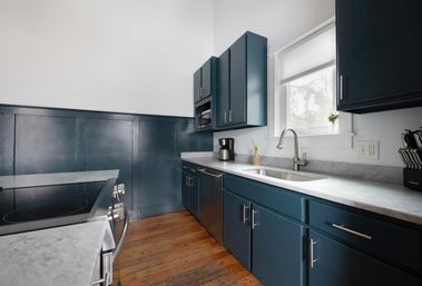 Bright kitchen interior with navy blue cabinets, white marble countertops, stainless steel sink and faucet, electric range, coffee maker, knife block, and warm hardwood floors under a window.
