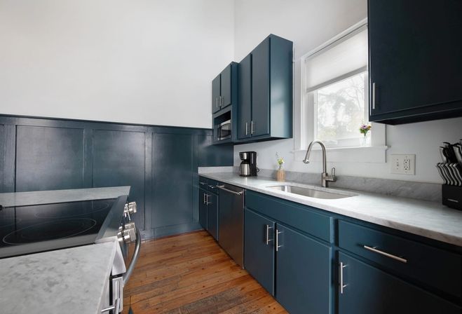 Bright kitchen interior with navy blue cabinets, white marble countertops, stainless steel sink and faucet, electric range, coffee maker, knife block, and warm hardwood floors under a window.