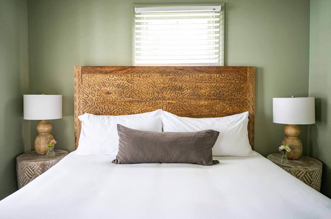 Cozy modern-rustic bedroom with carved wooden headboard, crisp white bedding, gray lumbar pillow, matching round nightstands with lamps and small vases against soft green walls.