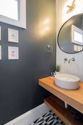 Stylish modern powder room with white round vessel sink on a floating wood vanity, large round mirror, wall-mounted faucet, dark gray accent wall with framed art, and geometric black-and-white floor tiles.