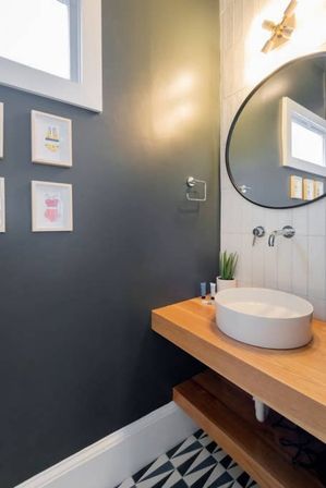 Stylish modern powder room with white round vessel sink on a floating wood vanity, large round mirror, wall-mounted faucet, dark gray accent wall with framed art, and geometric black-and-white floor tiles.