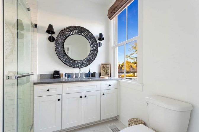 Bright, airy bathroom with white shaker vanity and gray countertop, round woven black mirror flanked by black sconces, glass shower door, toilet, and sunny window view of trees.