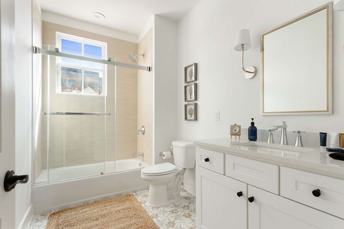 Bright modern residential bathroom with white shaker vanity and quartz countertop, framed mirror and sconce, toilet, tub with sliding glass doors and beige tile surround, patterned tile floor and jute rug bathed in natural light from window.
