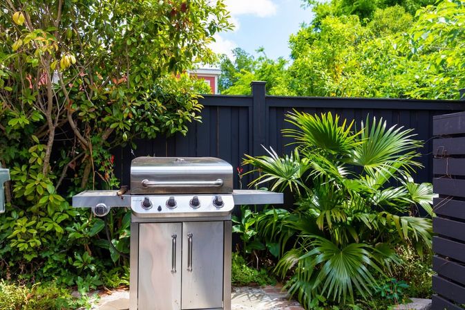 Stainless steel gas grill on a sunny backyard patio, surrounded by lush tropical palms and greenery in front of a black wooden fence, ready for a barbecue.
