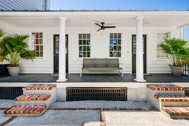 Sunlit white colonial-style front porch with two columns, brick steps, dark wood floor, metal bench, ceiling fan and potted palm plants — inviting coastal cottage entry.