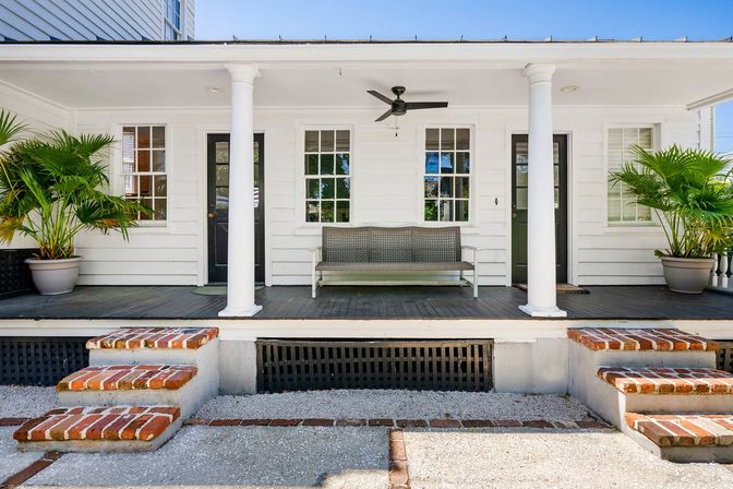 Sunlit white colonial-style front porch with two columns, brick steps, dark wood floor, metal bench, ceiling fan and potted palm plants — inviting coastal cottage entry.