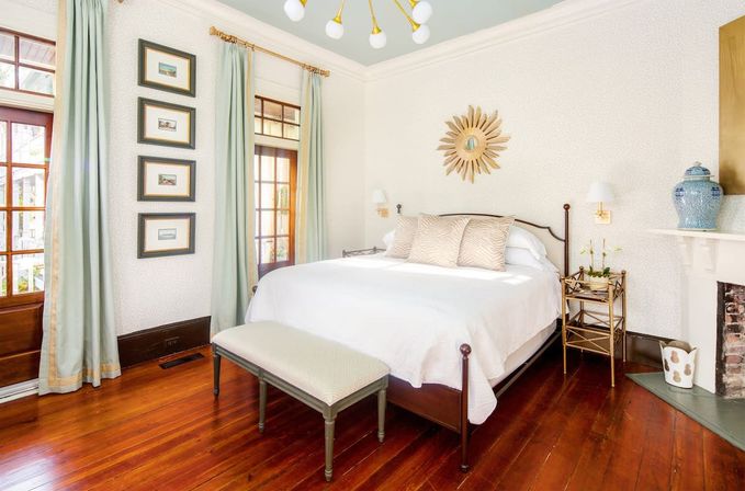 Bright airy bedroom with white bedding on a wooden bed frame, polished hardwood floors, pale green curtains over French doors, a sunburst wall mirror, brass bedside table and decorative fireplace.