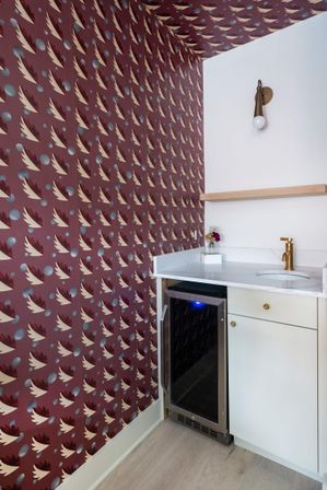 Cozy modern apartment wet bar corner with burgundy patterned wallpaper, white marble countertop, brass faucet, floating wood shelf, small vase of flowers and under-counter wine fridge glowing blue.