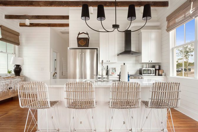 Sunny coastal farmhouse kitchen with white shiplap walls and exposed wooden beams, stainless steel fridge, white island with four woven rattan bar stools, black chandelier and large window with outdoor view.