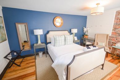 Cozy boutique guest bedroom with cream upholstered bed, blue accent wall and round mirror, matching bedside lamps, plaid pillows, exposed brick, hardwood floors, armchair and small desk