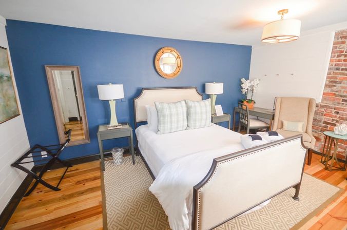 Cozy boutique guest bedroom with cream upholstered bed, blue accent wall and round mirror, matching bedside lamps, plaid pillows, exposed brick, hardwood floors, armchair and small desk
