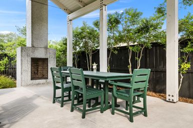Cheerful green outdoor dining set on a covered backyard patio with a concrete fireplace, small flower vase, white support posts and leafy trees.