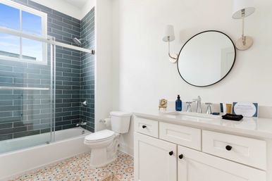 Bright modern bathroom with blue-gray subway-tiled shower and sliding glass door, white toilet and shaker-style vanity with round black mirror, wall sconces, and patterned tile floor.