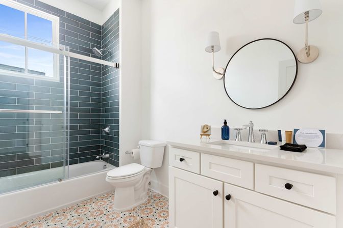 Bright modern bathroom with blue-gray subway-tiled shower and sliding glass door, white toilet and shaker-style vanity with round black mirror, wall sconces, and patterned tile floor.