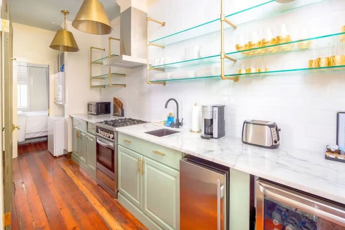 Bright galley kitchen with marble countertops, mint-green cabinets, brass accents and glass shelves, stainless appliances, wood floors and pendant lights.