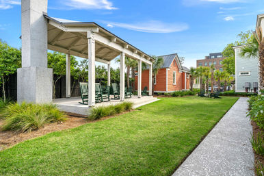 Sunny community courtyard with a covered pavilion and green rocking chairs overlooking a manicured lawn, palm trees, and an orange cottage-style building