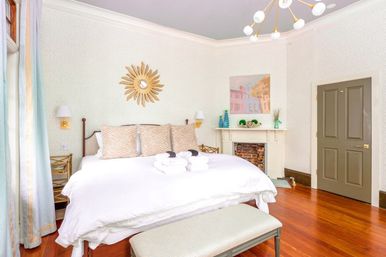 Bright airy guest bedroom with king-size bed in white linens, zebra-pattern pillows, sunburst mirror above the headboard, decorative fireplace mantel, hardwood floors and soft blue curtains.