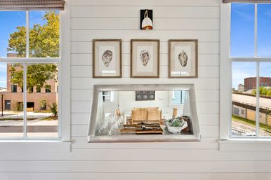 White shiplap wall with three framed oyster prints above a metal pass-through counter holding glassware and a decorative bowl, flanked by two windows with urban street and tree views.