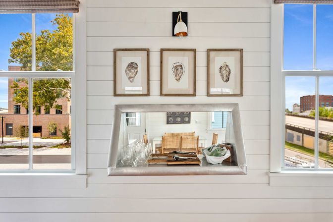 White shiplap wall with three framed oyster prints above a metal pass-through counter holding glassware and a decorative bowl, flanked by two windows with urban street and tree views.