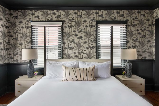 Stylish bedroom with white king bed centered between two windows, black-and-white tropical wallpaper featuring tall birds and palms, patterned ceramic bedside lamps on cream nightstands and a decorative lumbar pillow.