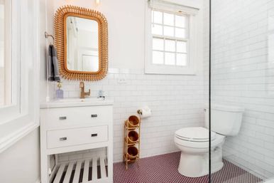 Bright modern bathroom with white subway tile walls, rattan-framed mirror above a white vanity, glass shower panel, toilet, and pink penny-tile floor — airy, coastal-inspired design.