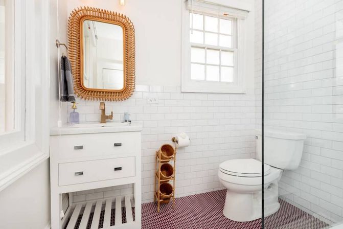 Bright modern bathroom with white subway tile walls, rattan-framed mirror above a white vanity, glass shower panel, toilet, and pink penny-tile floor — airy, coastal-inspired design.