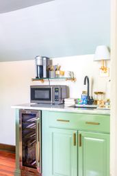 Cheerful mint-green kitchenette with marble countertop, microwave, single-serve coffee maker on a glass shelf, wine fridge, black faucet and brass hardware.