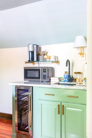 Cheerful mint-green kitchenette with marble countertop, microwave, single-serve coffee maker on a glass shelf, wine fridge, black faucet and brass hardware.