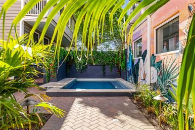 Compact urban plunge pool in a tropical courtyard framed by palm fronds and hanging vines, vibrant floral mural on the wall and brick paver patio — bright relaxing backyard oasis