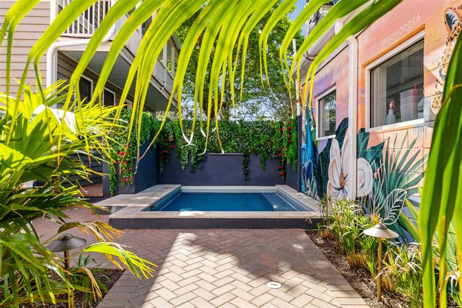 Compact urban plunge pool in a tropical courtyard framed by palm fronds and hanging vines, vibrant floral mural on the wall and brick paver patio — bright relaxing backyard oasis