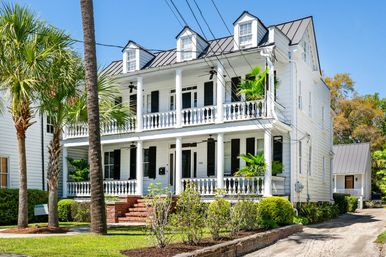 White historic Southern two-story house with stacked porches, classical columns, black shutters and a metal roof, framed by palm trees and manicured shrubs on a sunny day