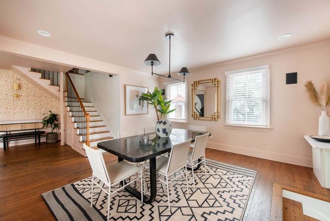 Bright modern dining room interior with a black marble table and white chairs on a black-and-white geometric rug, pendant lights overhead, gold-framed mirror, potted plant, wooden floors and a staircase in the background.