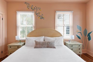 Sunlit peach bedroom with rattan headboard, crisp white bedding and lumbar pillow, two windows with white blinds, matching vintage nightstands with lamps and flowers, blue botanical wall decals and hardwood floor.