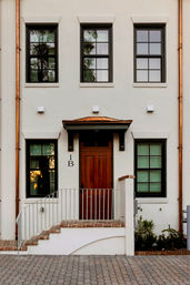Charming white stucco townhouse entrance with a rich wooden door, symmetrical dark-framed windows, copper downspouts, brick steps and metal railing opening onto a cobblestone driveway.