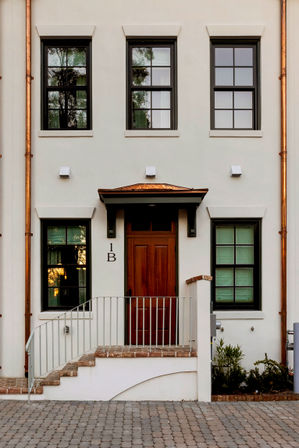 Charming white stucco townhouse entrance with a rich wooden door, symmetrical dark-framed windows, copper downspouts, brick steps and metal railing opening onto a cobblestone driveway.