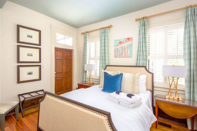Sunlit coastal-style guest bedroom with upholstered bed, blue accent pillow, folded towels, plaid curtains, plantation shutters, wooden door and bedside lamps.